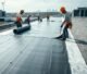 Roofing workers installing waterproof membrane during commercial flat roof replacement on a large building.